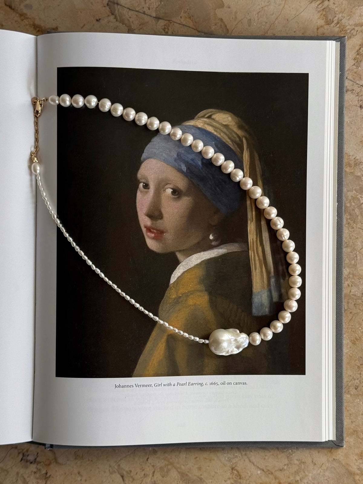 A pearl 14k gold-filled necklace draped over an open book, with a portrait of Vermeer's Girl with a Pearl Earring printed on one of the pages. The book lies on a textured beige stone surface.