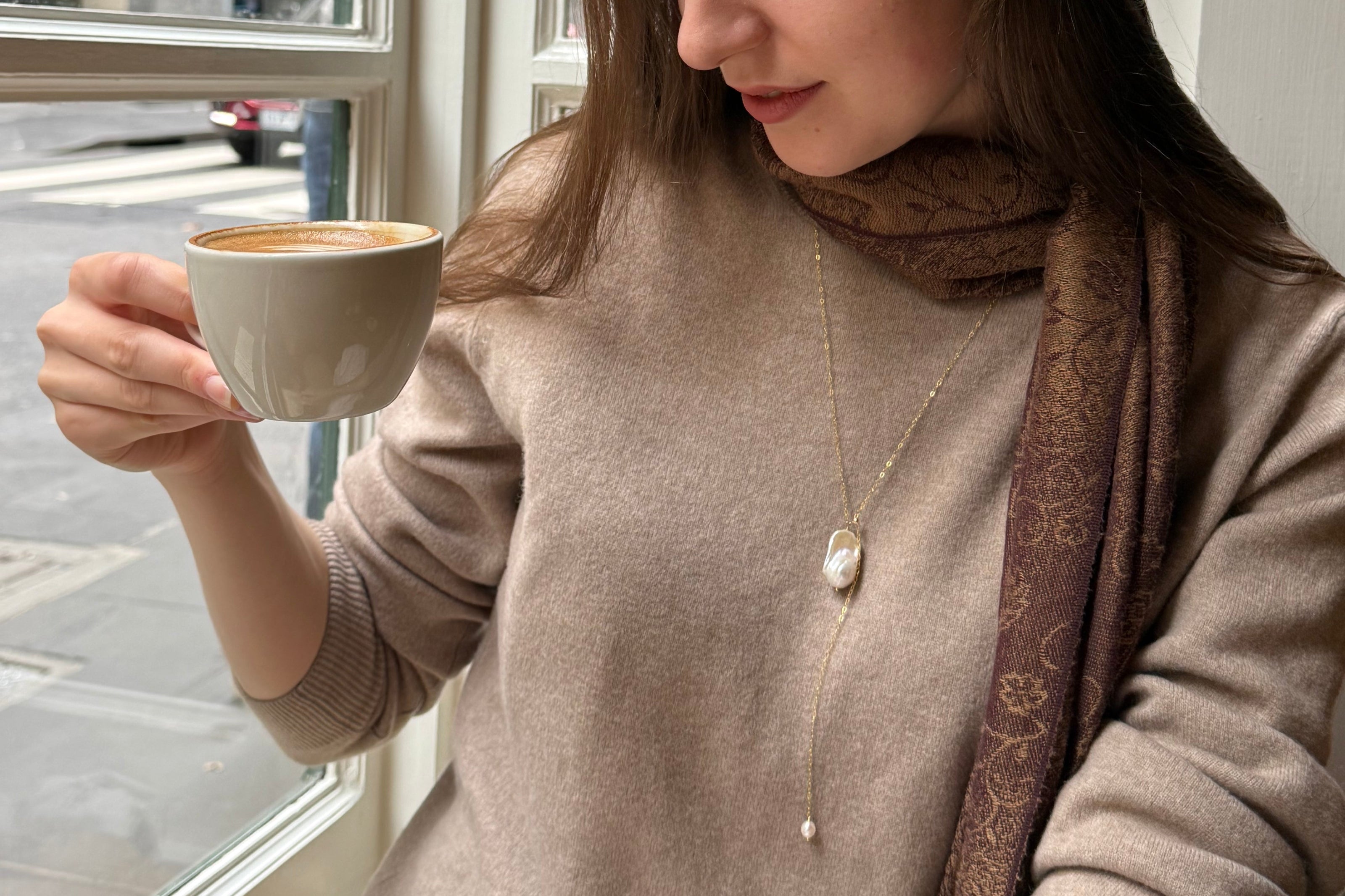 Woman holding a cup of coffee wearing a baroque pearl necklace and scarf, sitting on a couch.