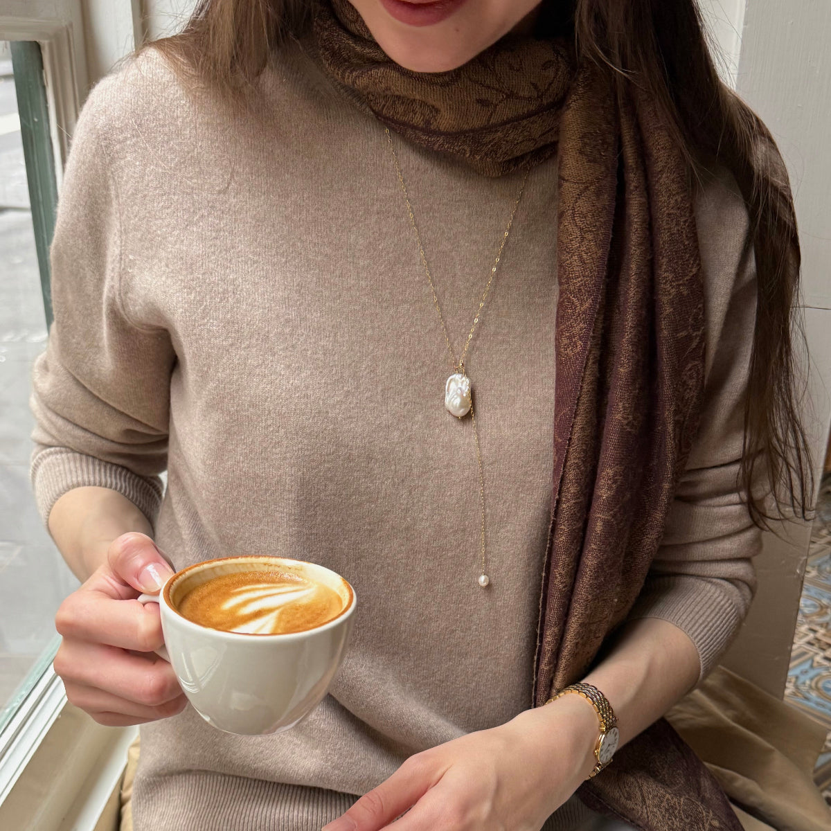 Person holding a cup of coffee wearing a baroque pearl necklace and scarf, sitting on a couch.