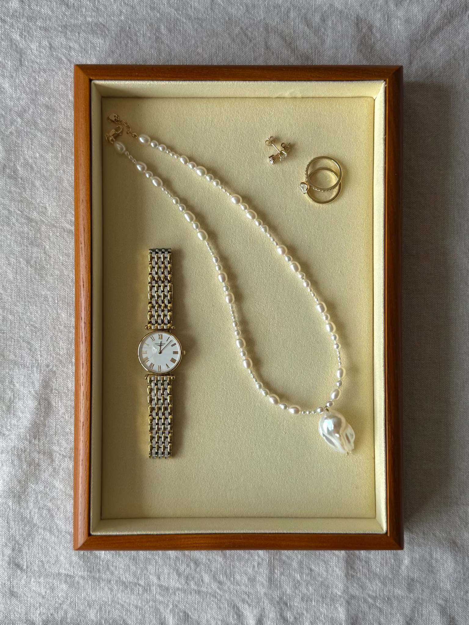 A curated jewelry and watch display inside a rectangular wooden presentation box with a cream velvet lining, placed on a textured light gray fabric background. The collection includes a long pearl necklace with a white irregular pendant, a gold-tone watch with a silver-tone mesh bracelet, two gold rings, and a small gold stud earring.