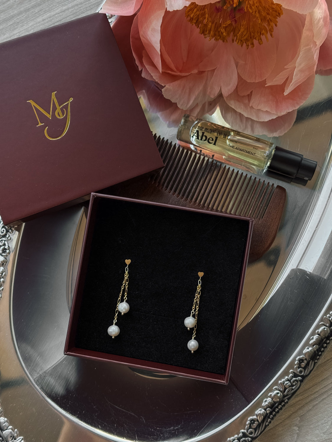 Gold earrings with pearls in a box on a reflective surface with a flower and perfume bottle in the background.