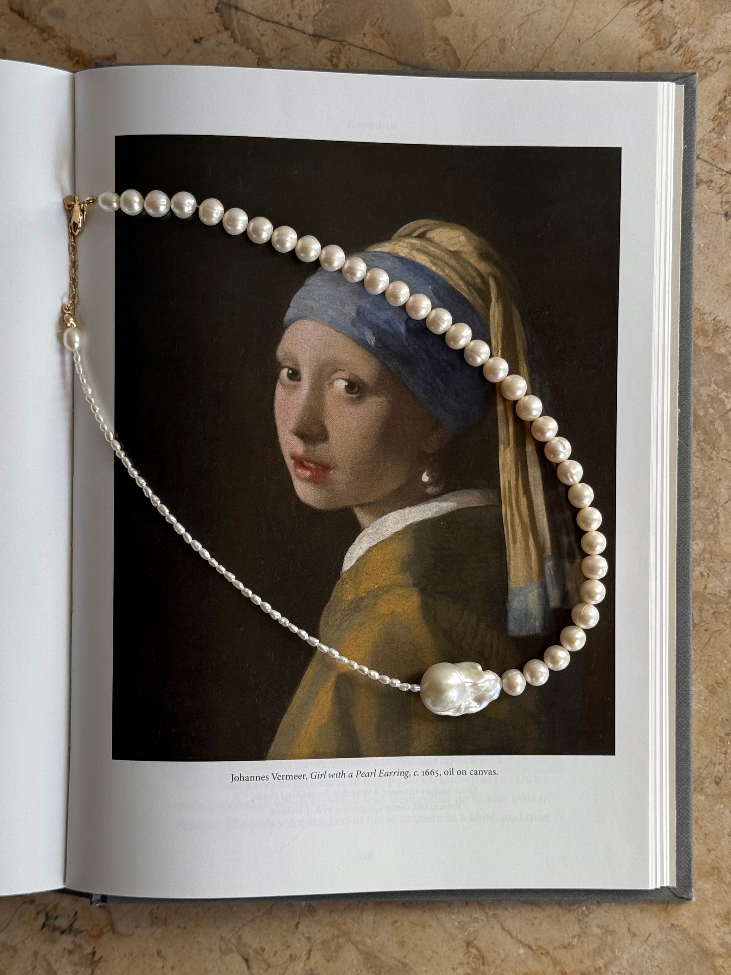 A pearl 14k gold-filled necklace draped over an open book, with a portrait of Vermeer's Girl with a Pearl Earring printed on one of the pages. The book lies on a textured beige stone surface.