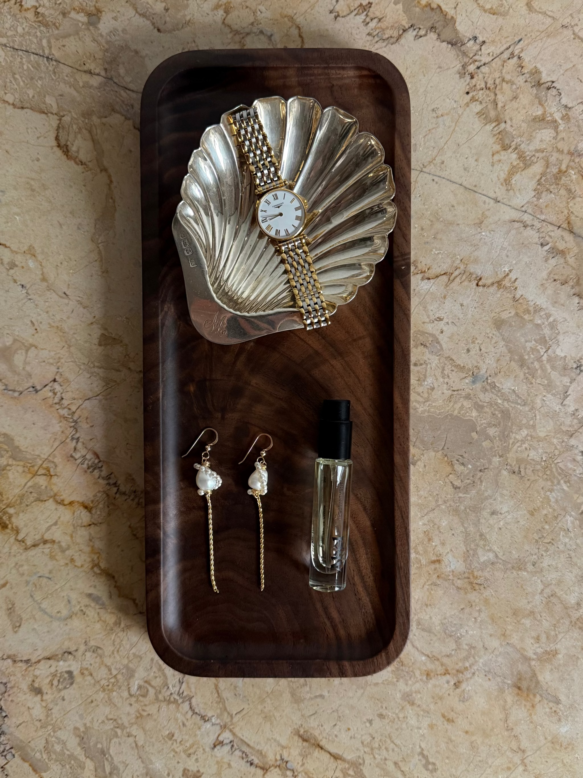 A styled flat-lay featuring a gold-tone watch with a textured shell-shaped bezel and round white dial, displayed on a dark wood tray alongside matching 14k gold filled dangle earrings with white pearl drops and a small clear glass perfume bottle, all set on a beige marble surface.