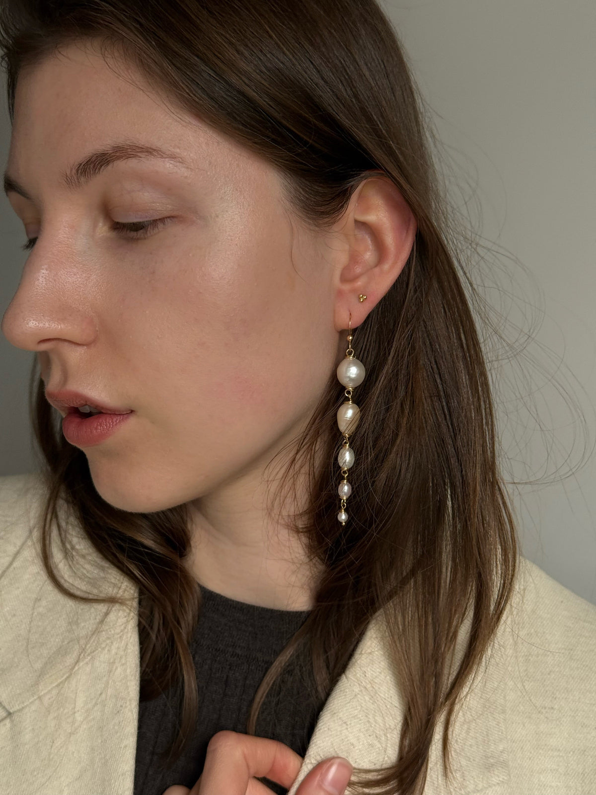 A close-up of a woman wearing a long, multi-tiered dangle earring made of cream-colored pearls and 14k gold filled hooks, set against a neutral gray background.